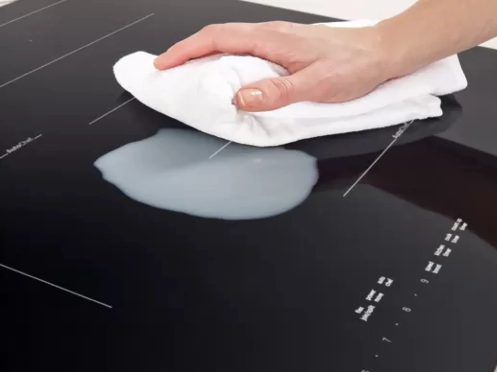 Hand wiping a spilled liquid on a black glass stovetop with a white cloth.