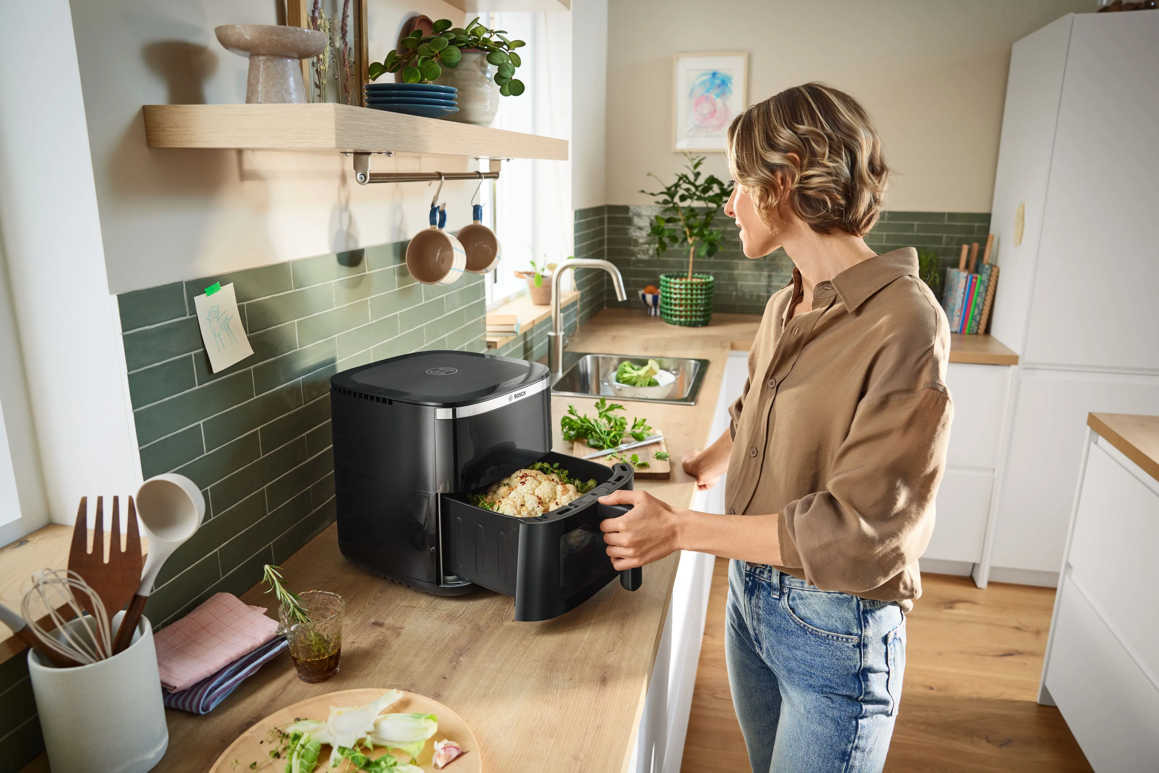 A young man slides a drawer filled with raw food into his Bosch Air Fryer, preparing for cooking in a bright kitchen.