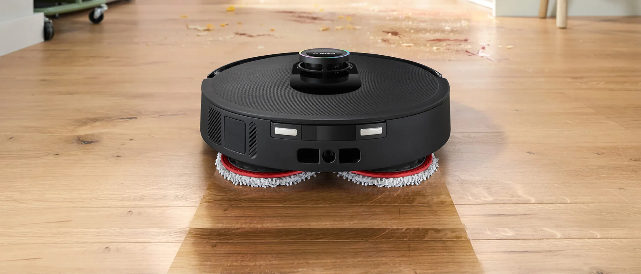 Robotski sesalnik sesa tla v kuhinji, na katerih je razlita hrana, medtem ko družina v ozadju pripravlja hrano.