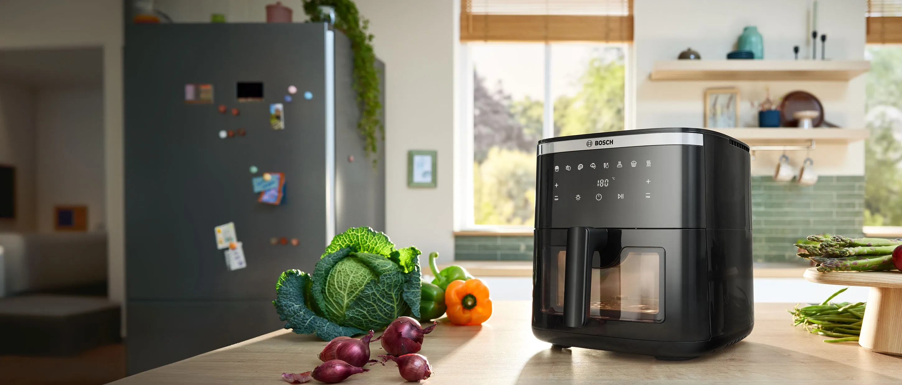 Two Bosch Air Fryer models positioned side by side on a bright kitchen counter. Several plates and bowls of food, including herb bread, chicken, roast, and fries, are placed next to the appliances, showcasing dishes made using the Air Fryers.
