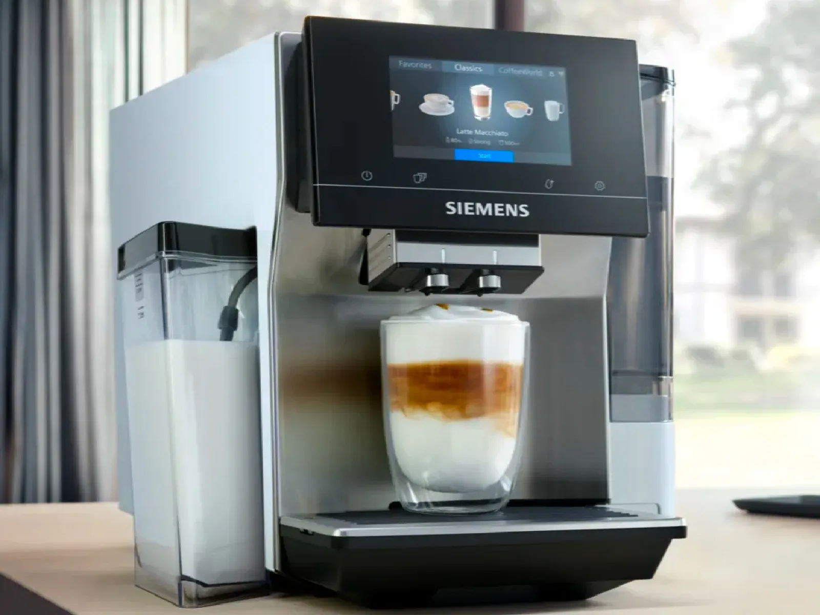 Siemens kafijas automāts ar skārienekrānu, kas pasniedz latte glāzi uz galda pie loga.