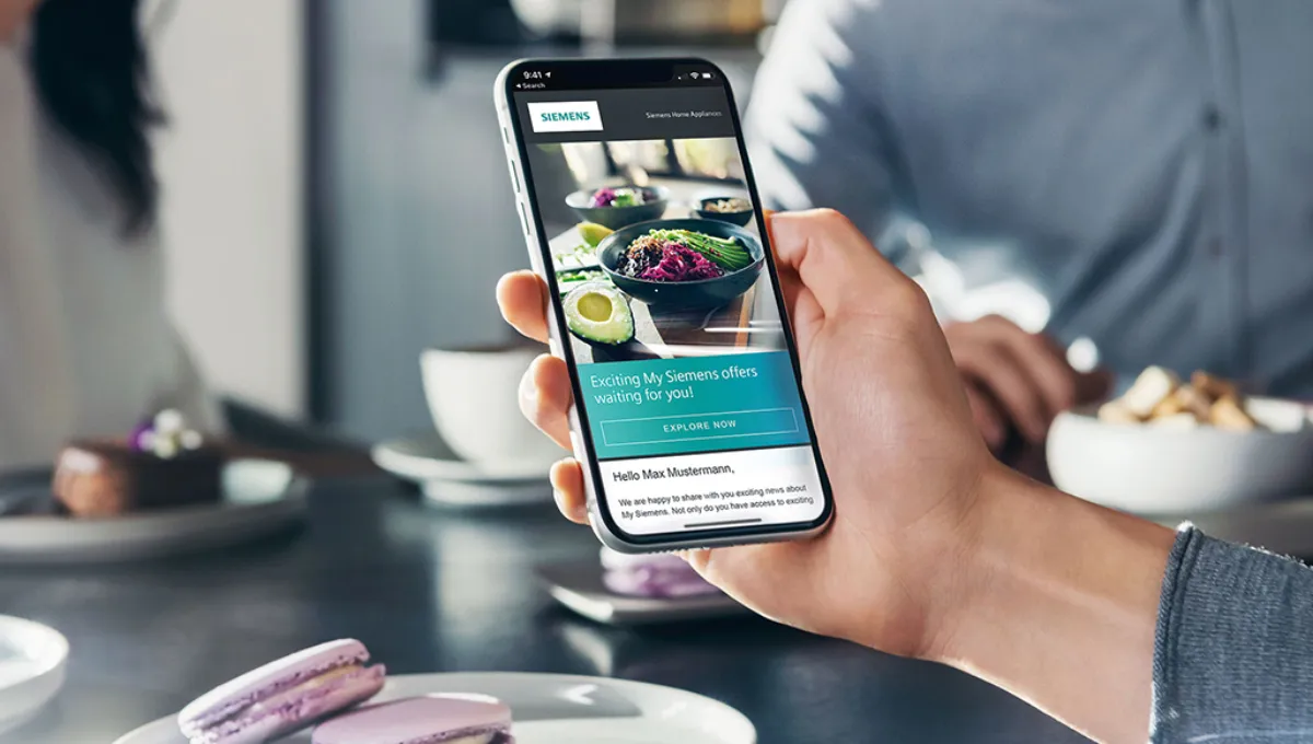 Hånd der holder en smartphone, som viser Siemens' hjemmeside, med lilla makroner på en tallerken og slørede personer i baggrunden ved et spisebord.