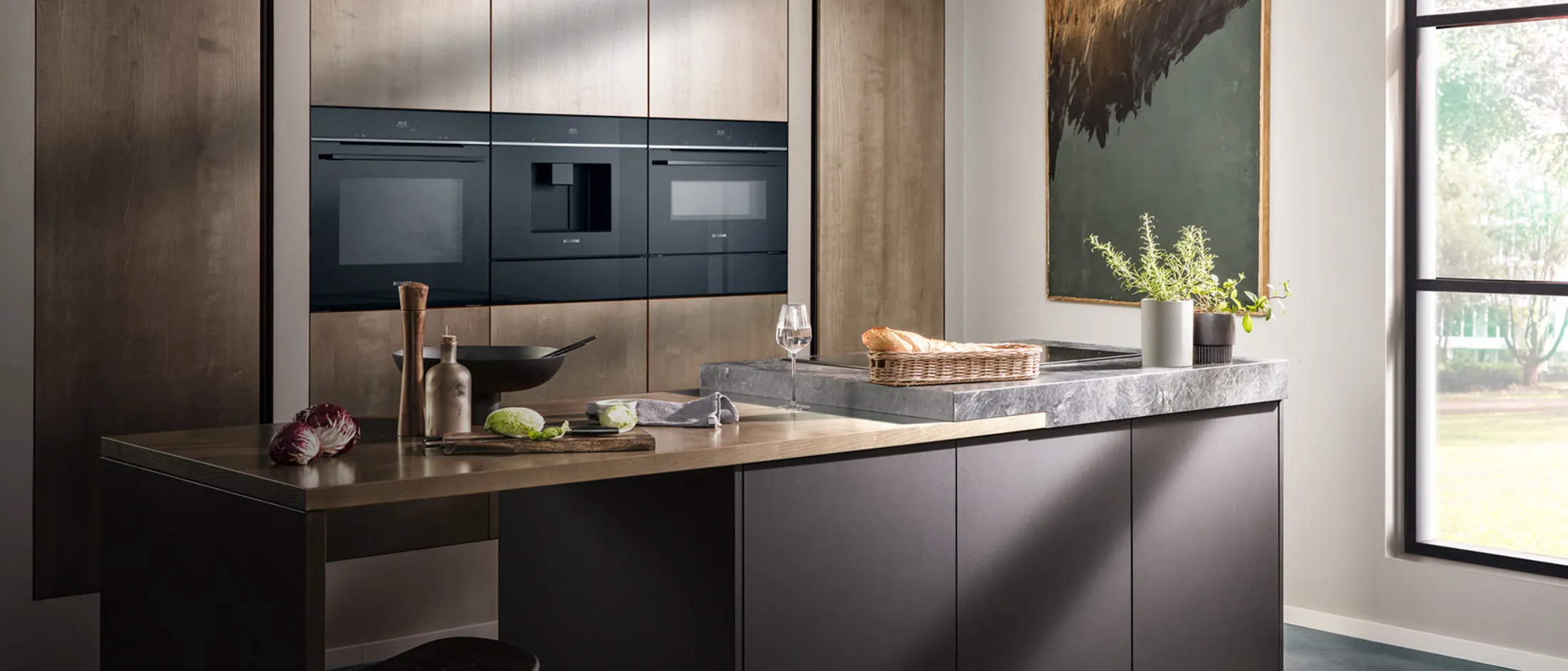 Modern kitchen featuring sleek built-in black appliances, a stone countertop, and decorative elements like a painting and a bread basket. 