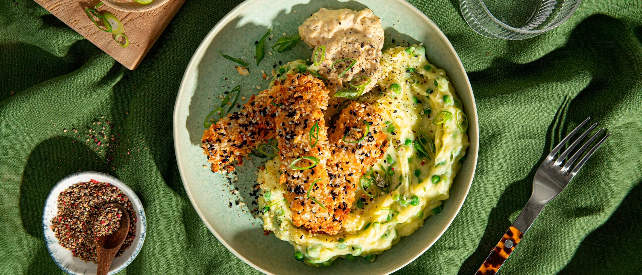 Plate of crusted chicken with mashed potatoes, peas, and sauce on a green cloth background.