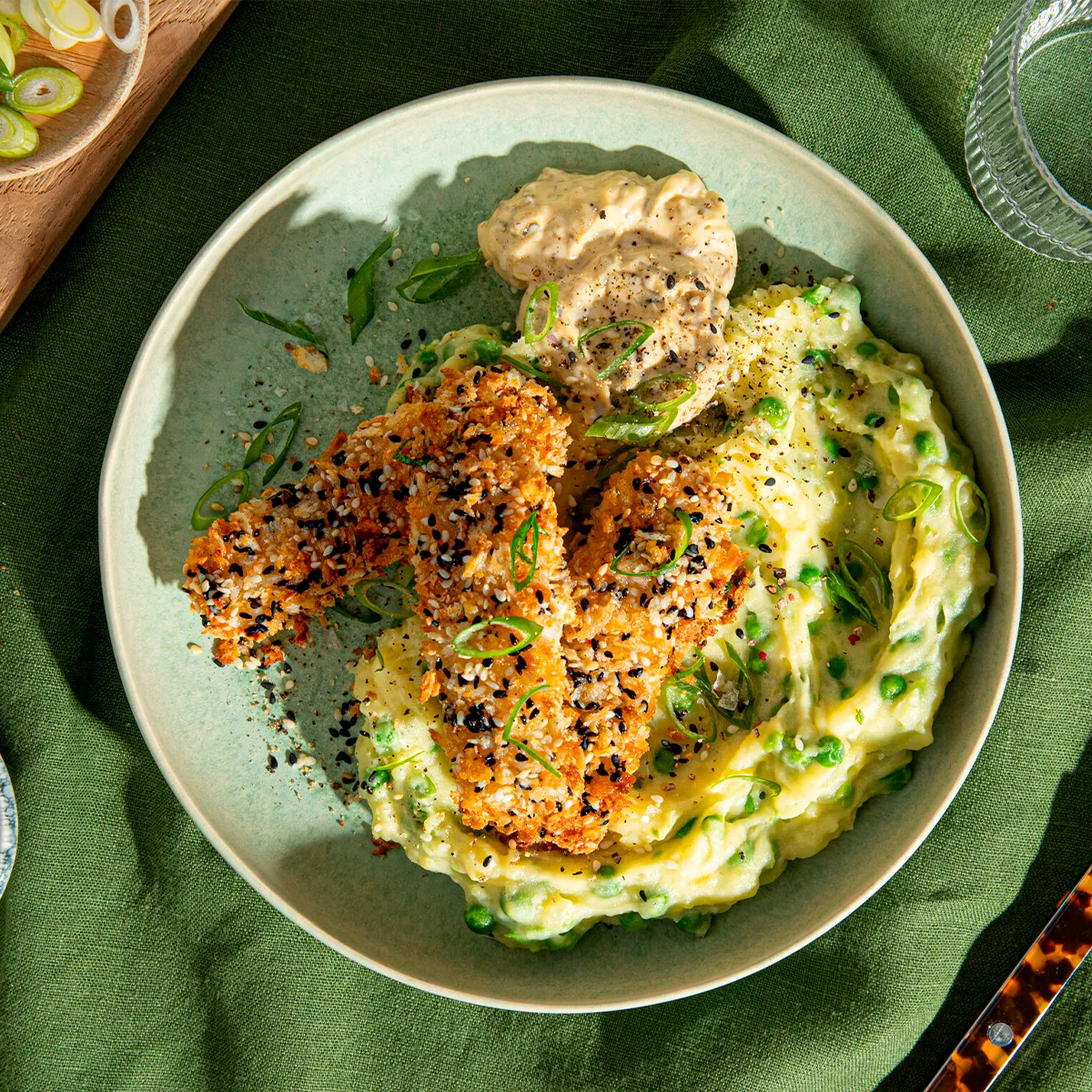 Plate of crusted chicken with mashed potatoes, peas, and sauce on a green cloth background.