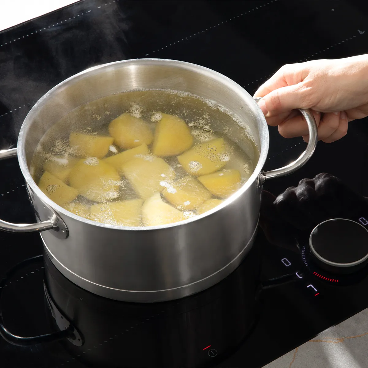 Hand holding a pot of boiling water with potatoes on a black induction cooktop.