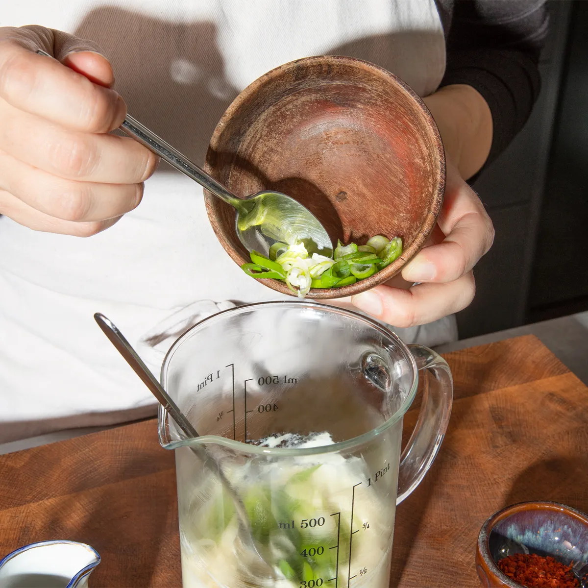 Person adding green onions from a wooden bowl into a glass measuring pitcher with other ingredients.