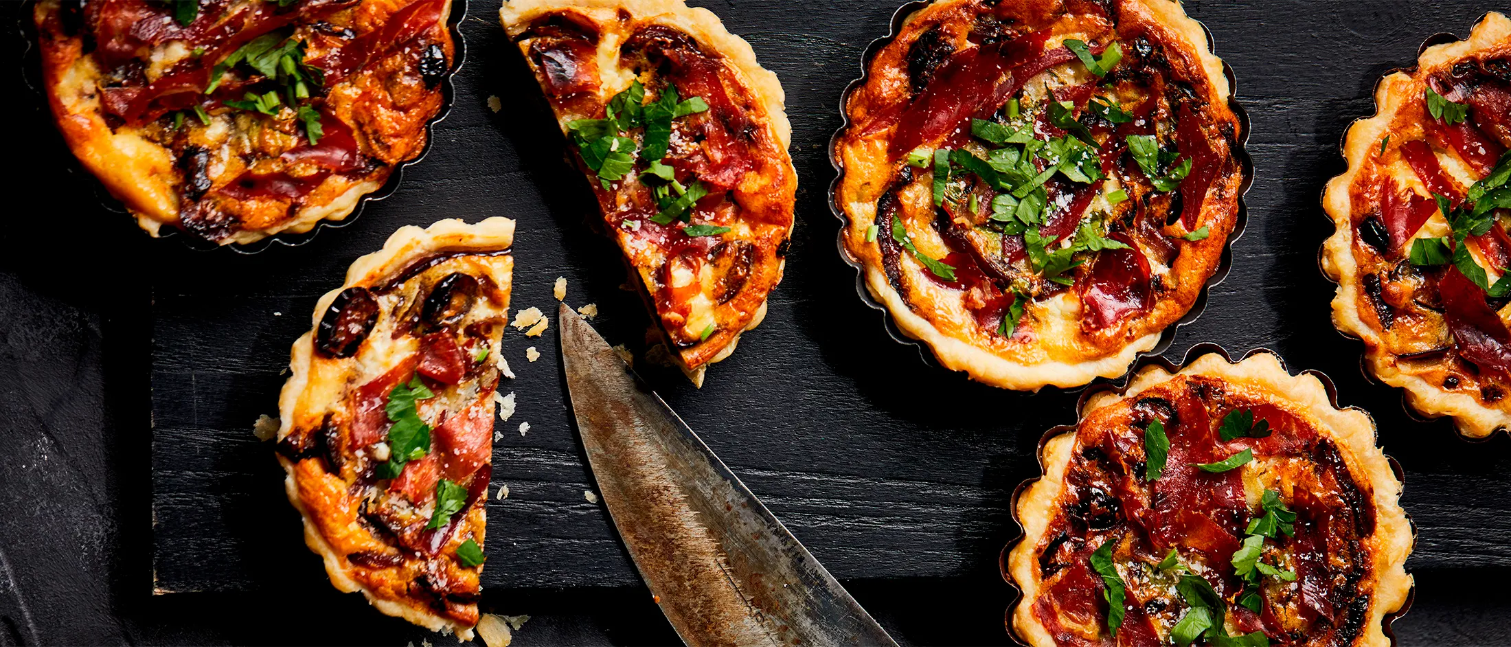 Five savory tarts with herbs and pepperoni displayed on a dark slate surface with a knife.