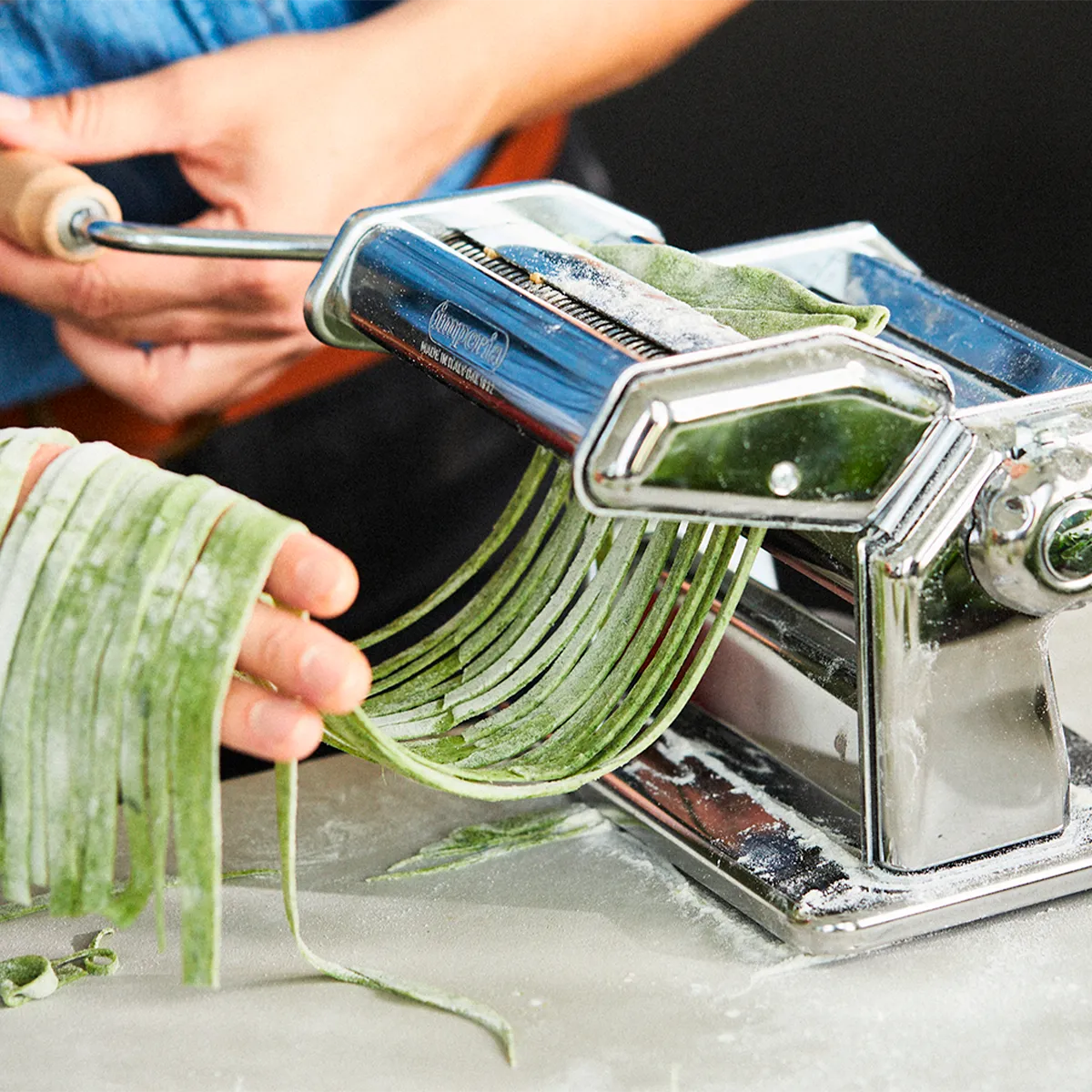 A person using a metal pasta machine to make green fettuccine. The machine is on a floured surface.