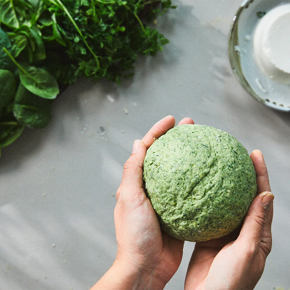 Hands holding a ball of green dough with fresh greens and a plate in the background.