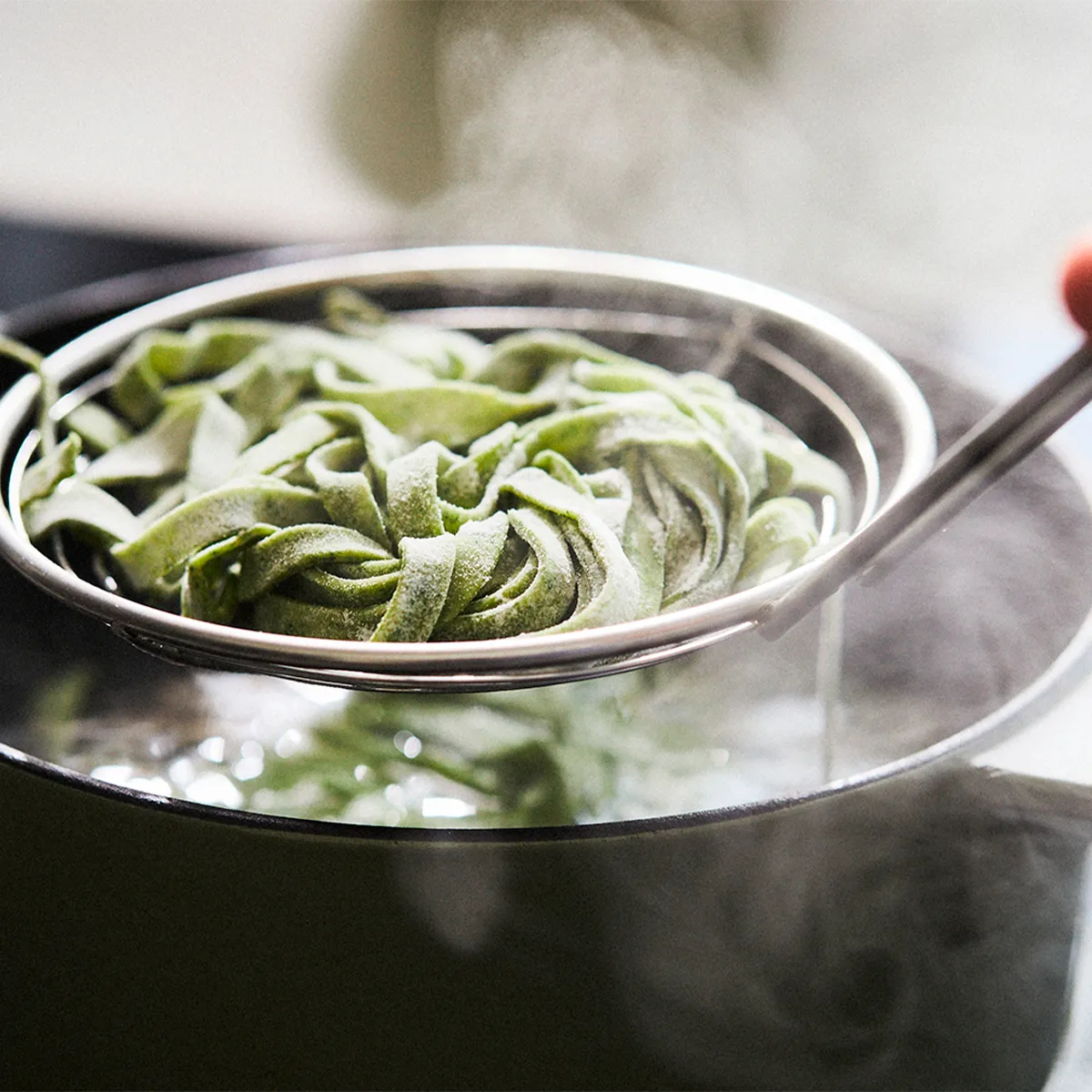 A metal strainer with frozen green pasta held over a pot of boiling water with steam rising.
