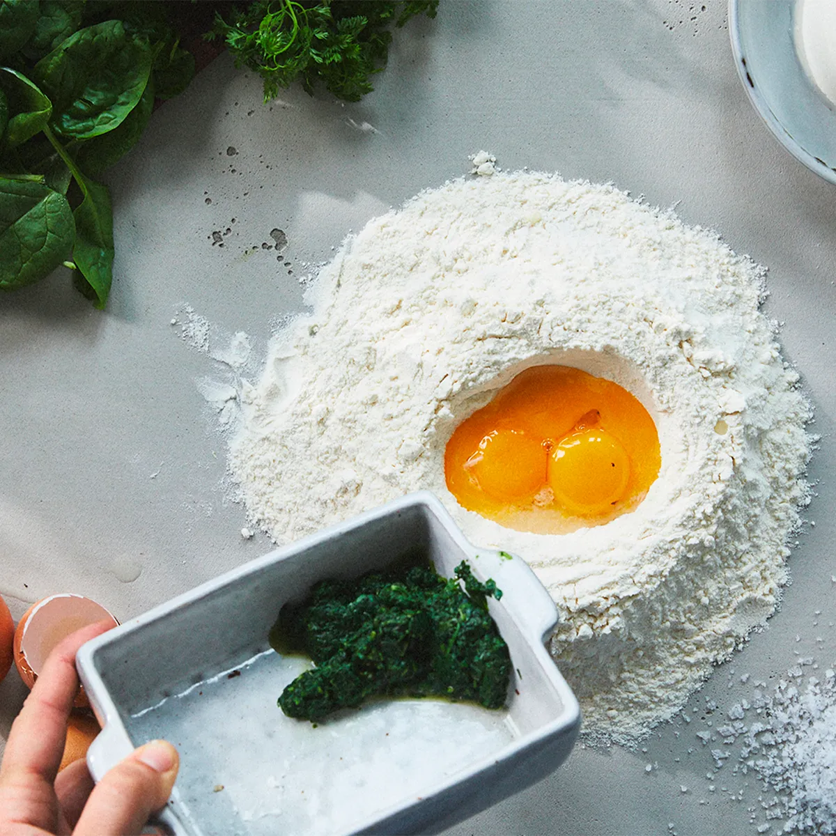 Flour mound with cracked eggs in center, spinach held in a dish nearby, ready for mixing.