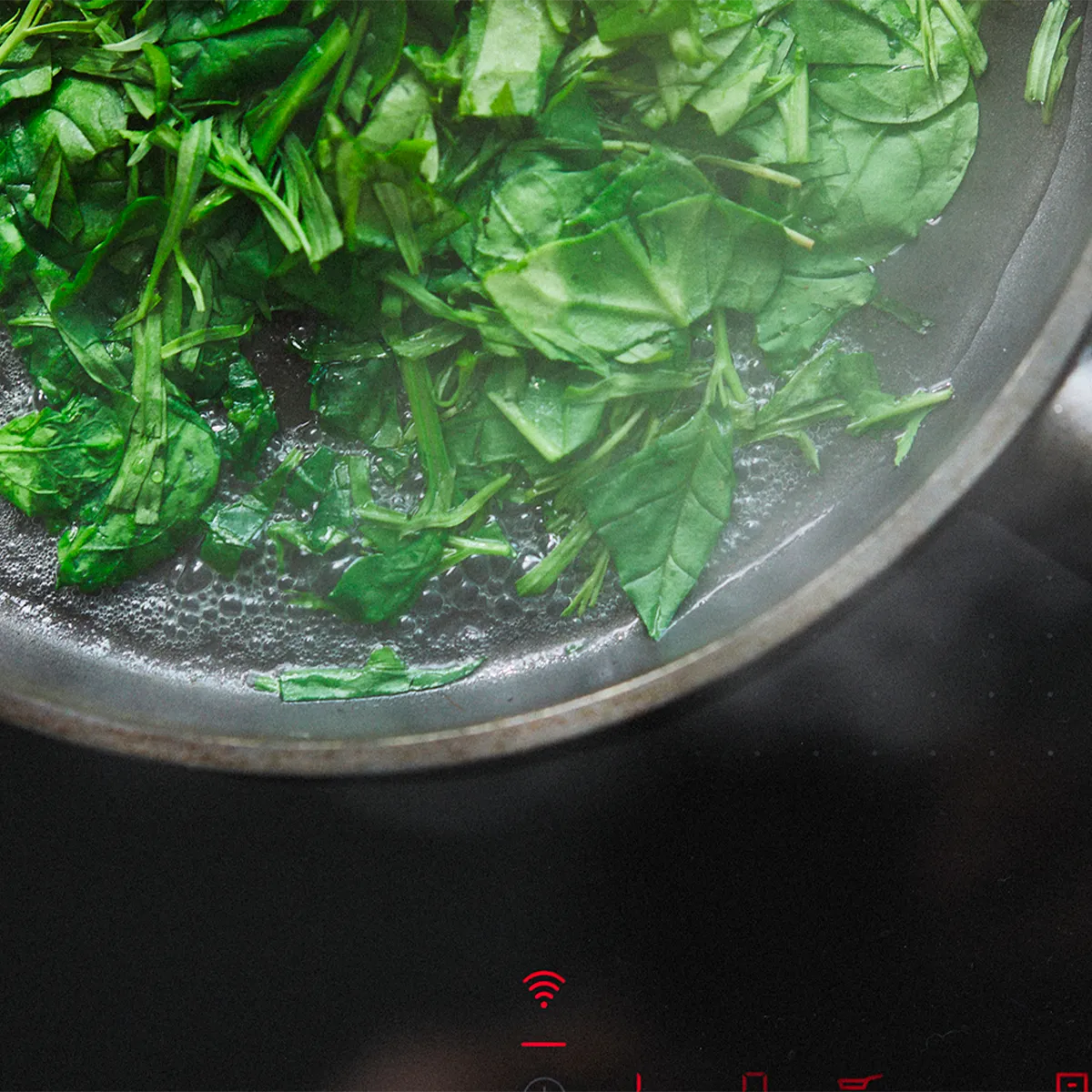 Fresh spinach leaves being sautéed in a frying pan on a stove with visible steam.