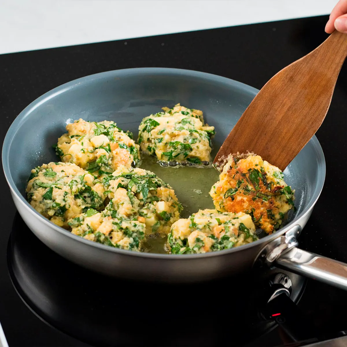 A frying pan with vegetable patties being cooked on a stovetop, wooden spatula flipping one.
