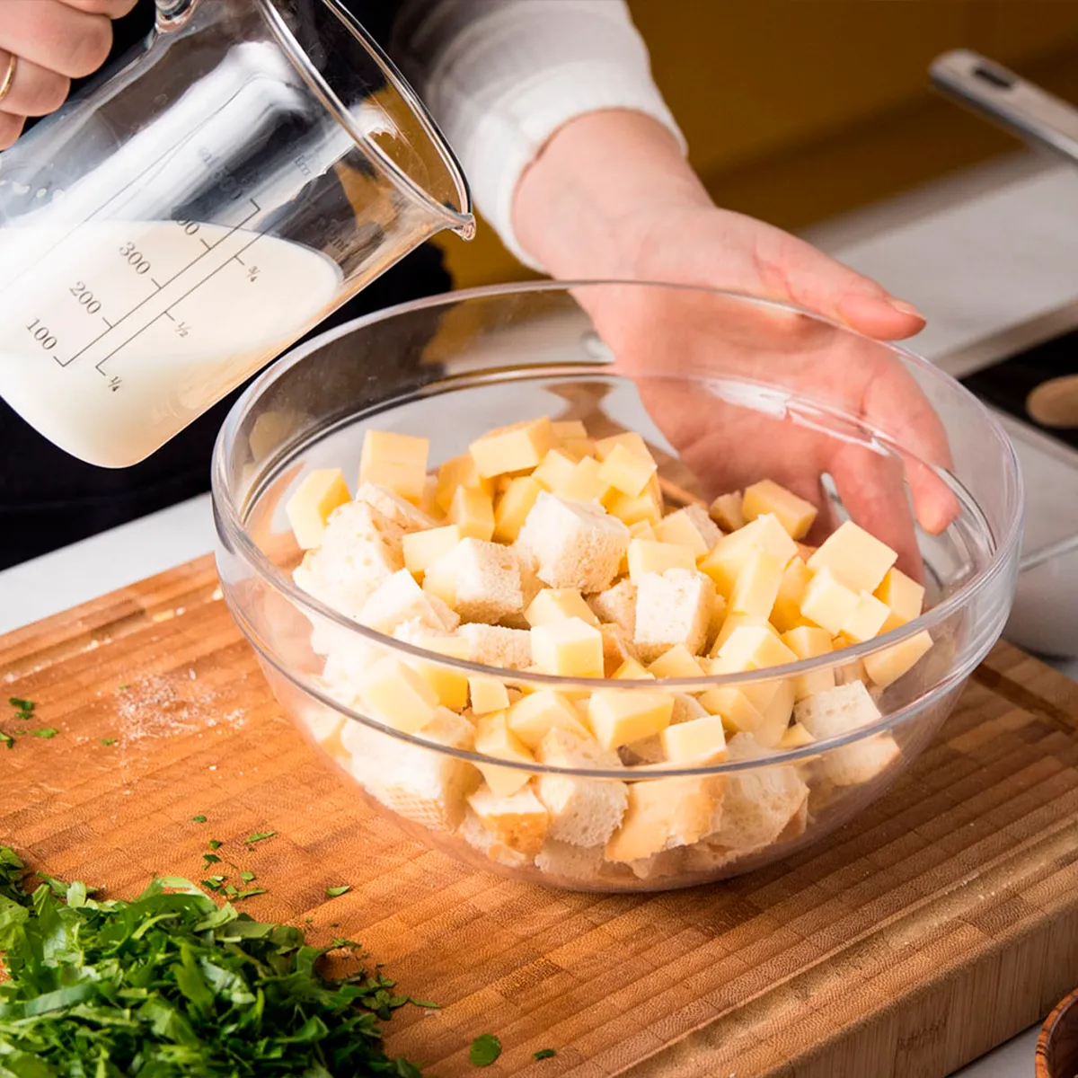 Person pouring milk into a bowl of bread and cheese cubes on a wooden cutting board.