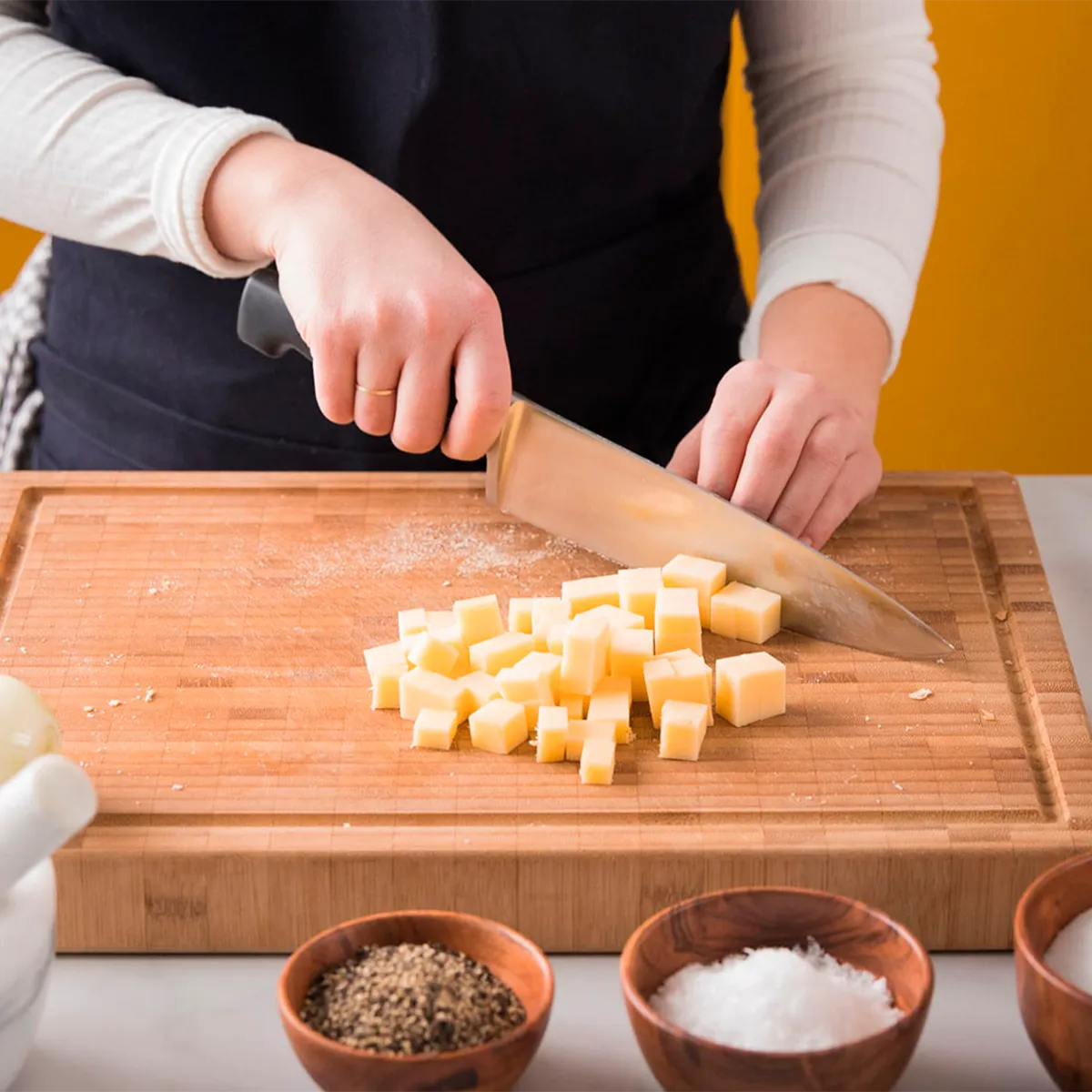 Person slicing cheese into cubes on a wooden cutting board, with spices in small bowls nearby.