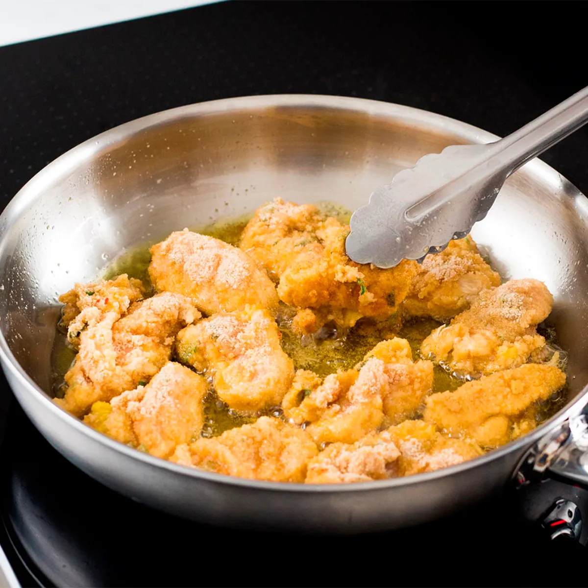 Frying pan with pieces of breaded chicken being cooked in hot oil, held by metal tongs.