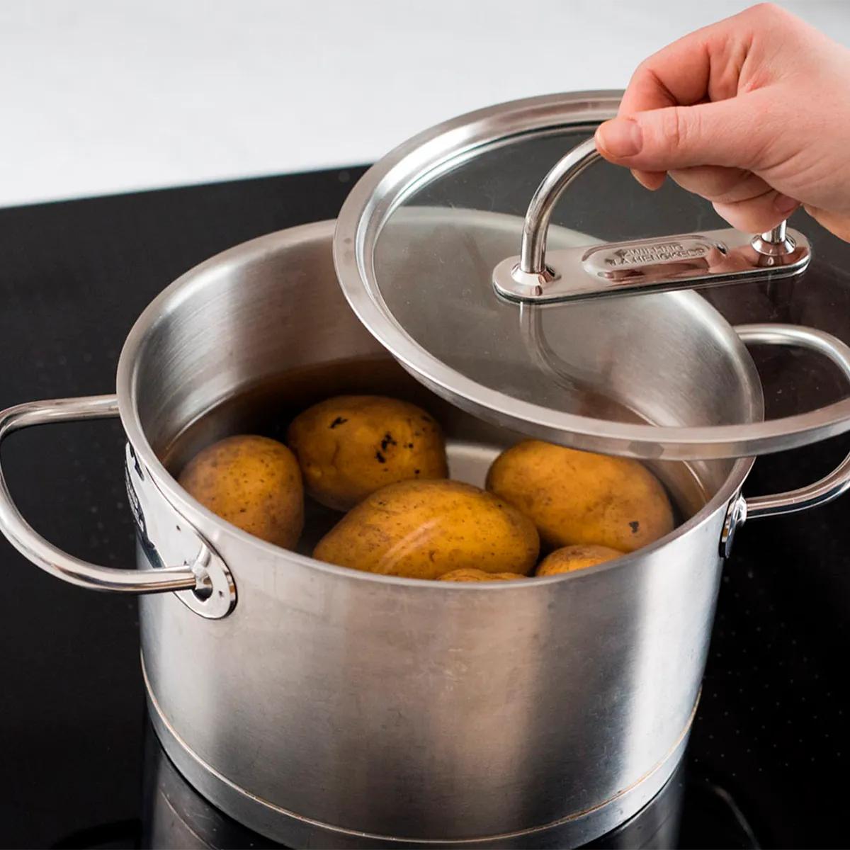 A hand lifting the lid of a metal pot with potatoes boiling in water on a stovetop.