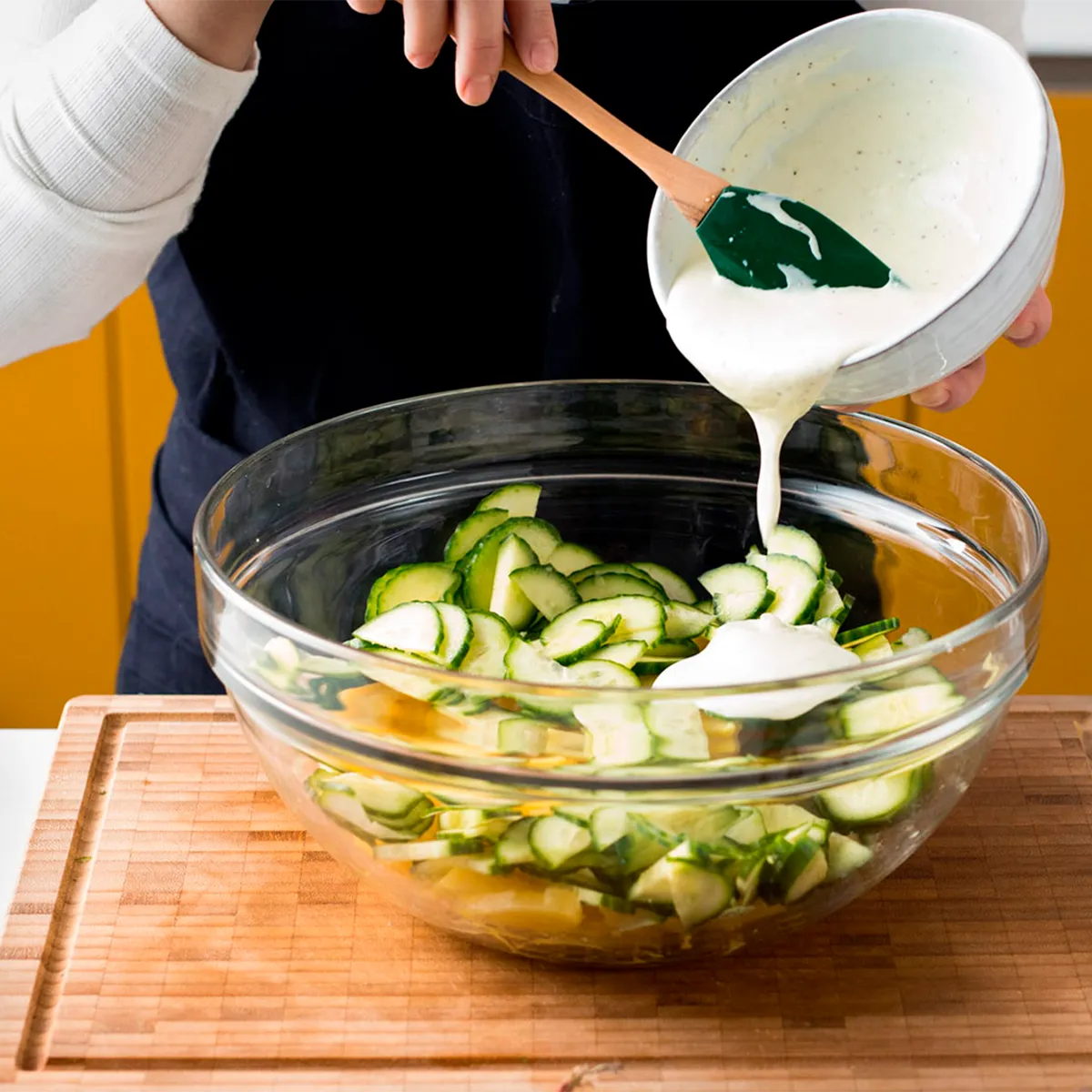 Person pouring creamy dressing onto sliced cucumbers in a glass bowl on a wooden cutting board.