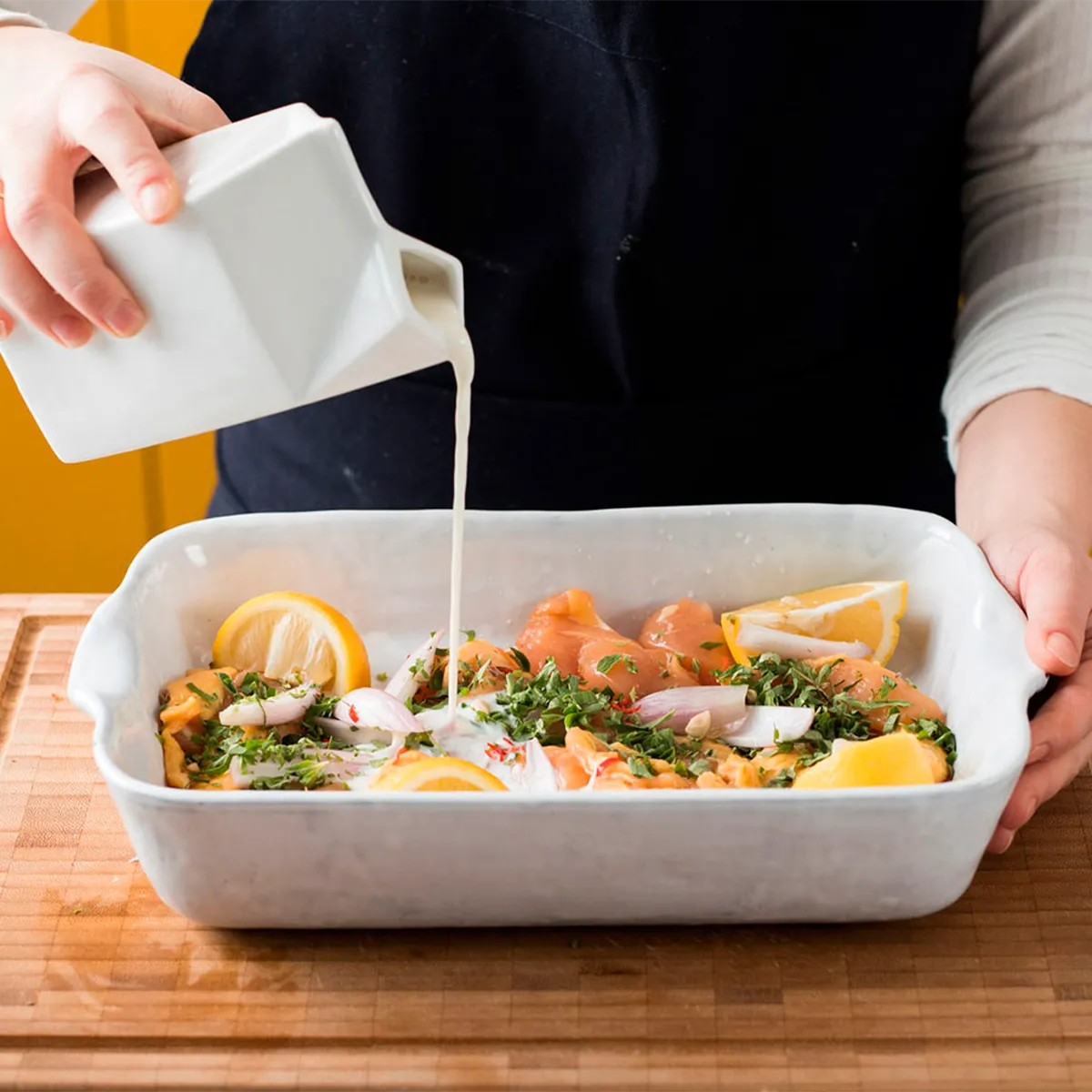 Person pouring white liquid over marinated chicken and lemon slices in a white baking dish.