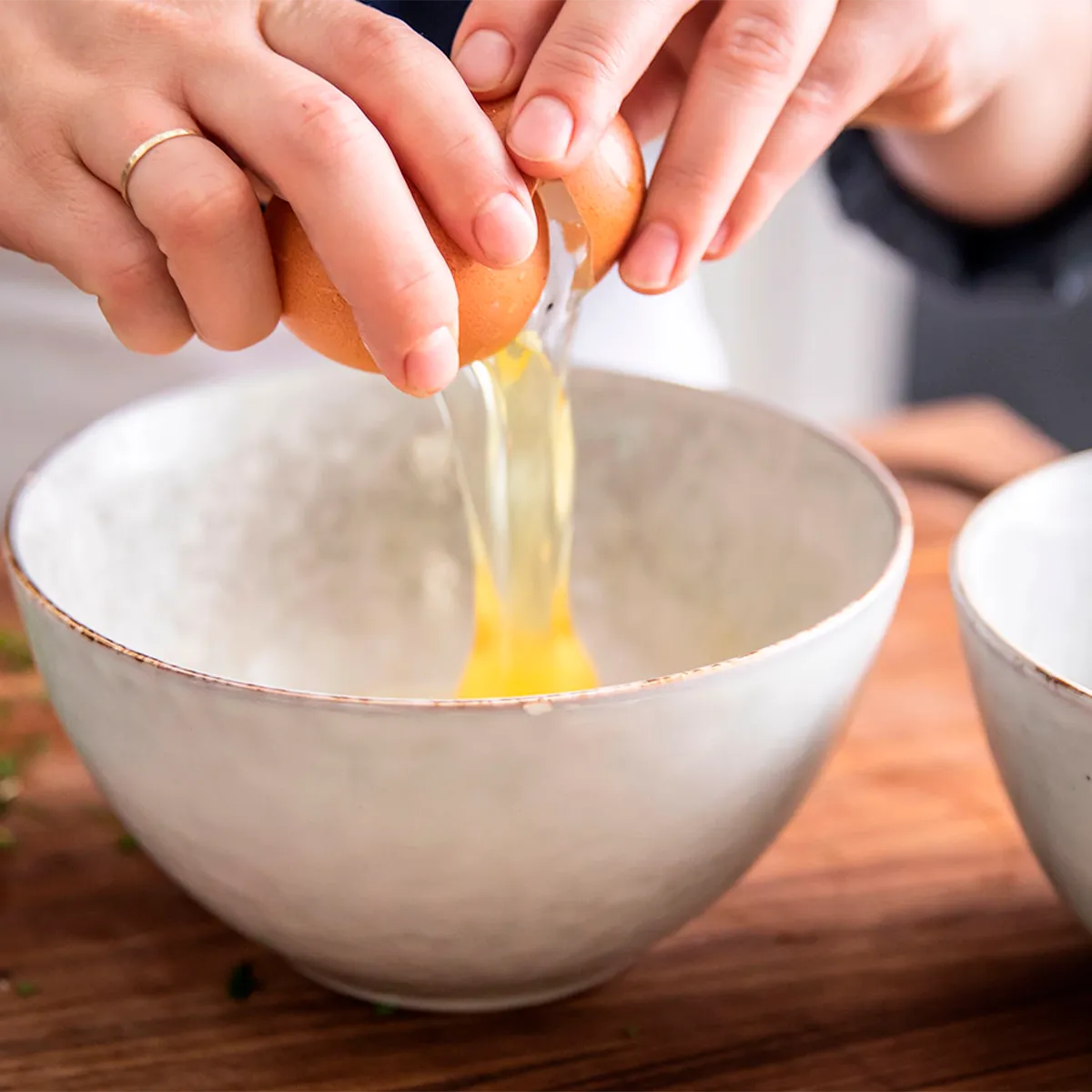 Hands cracking an egg into a large white ceramic bowl on a wooden countertop.