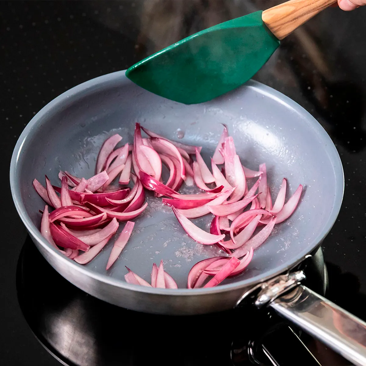 A non-stick frying pan with sliced red onions being stirred with a green spatula on a stovetop.