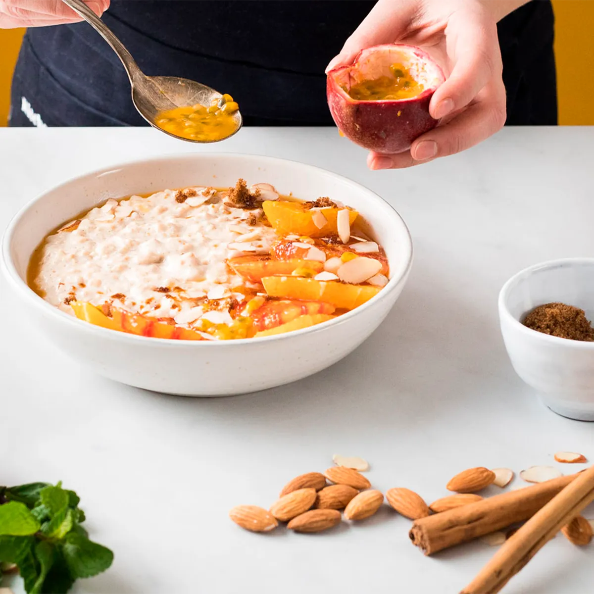 Person adding passion fruit to a bowl of oatmeal garnished with fruit slices and nuts.