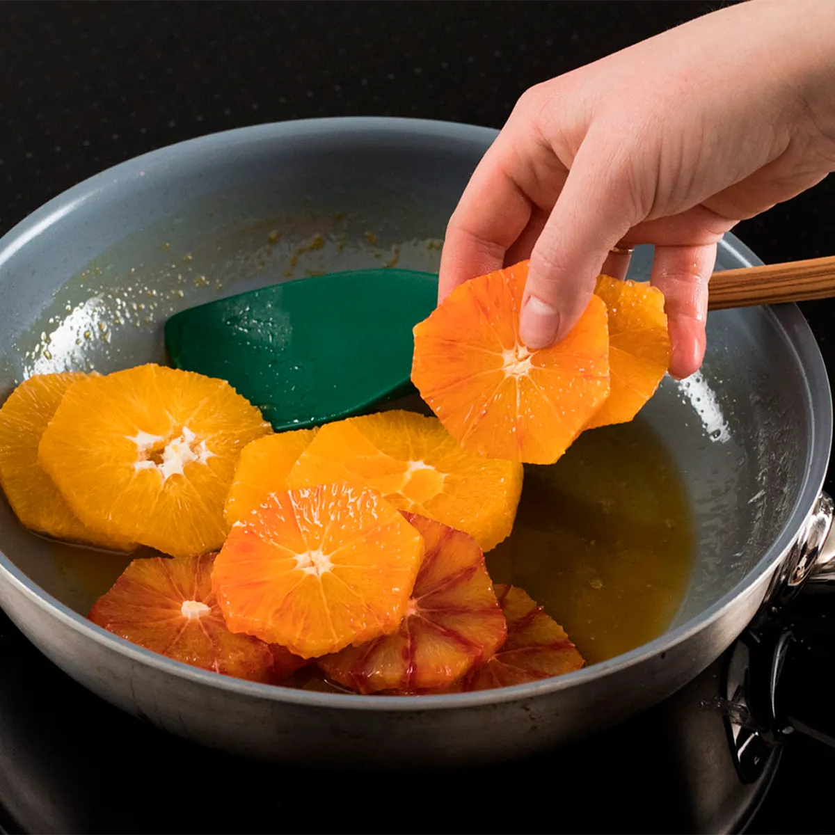 Hand placing orange slices into a frying pan with a green spatula, cooking in oil.