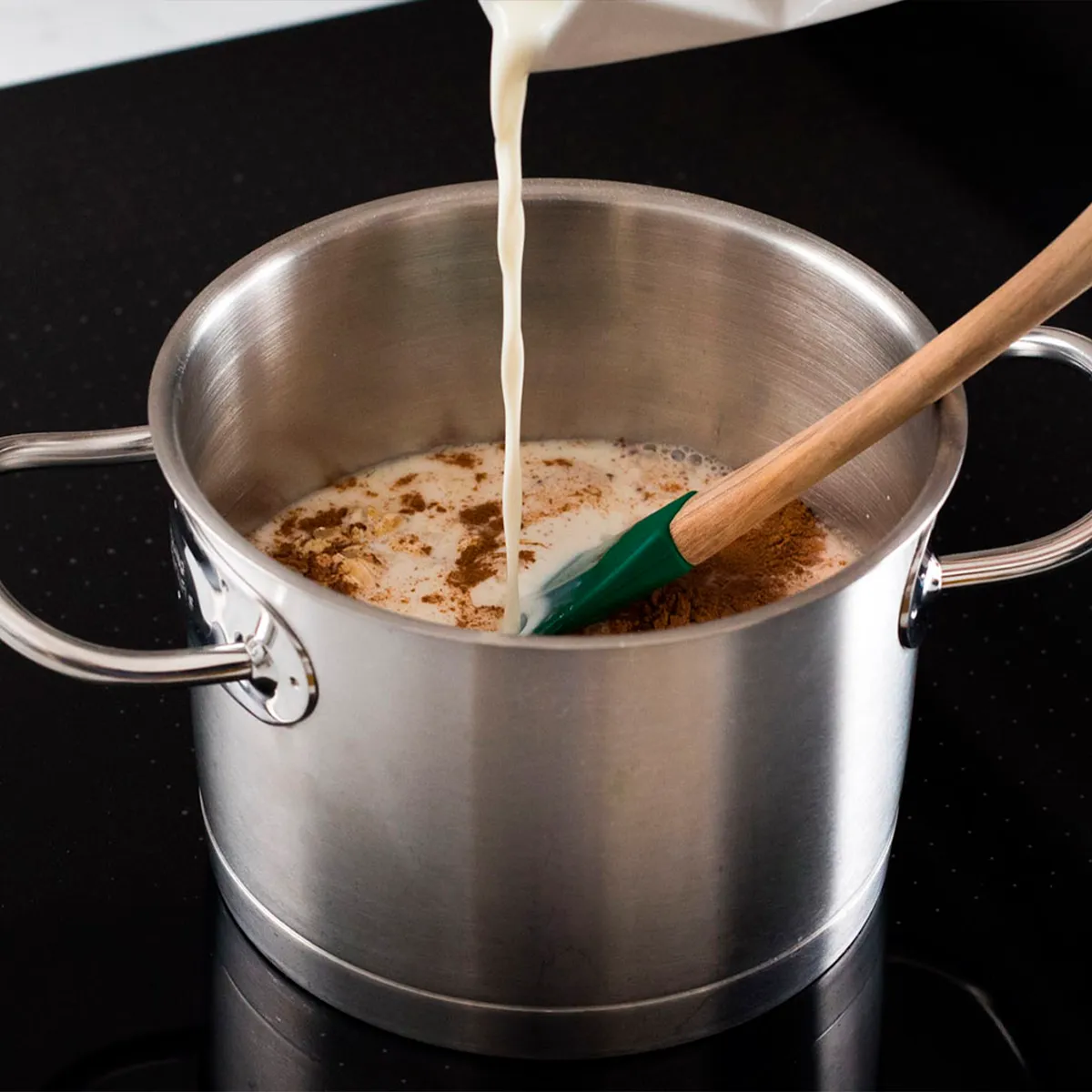 Milk is being poured into a stainless steel pot on a stove, with a wooden spoon inside.