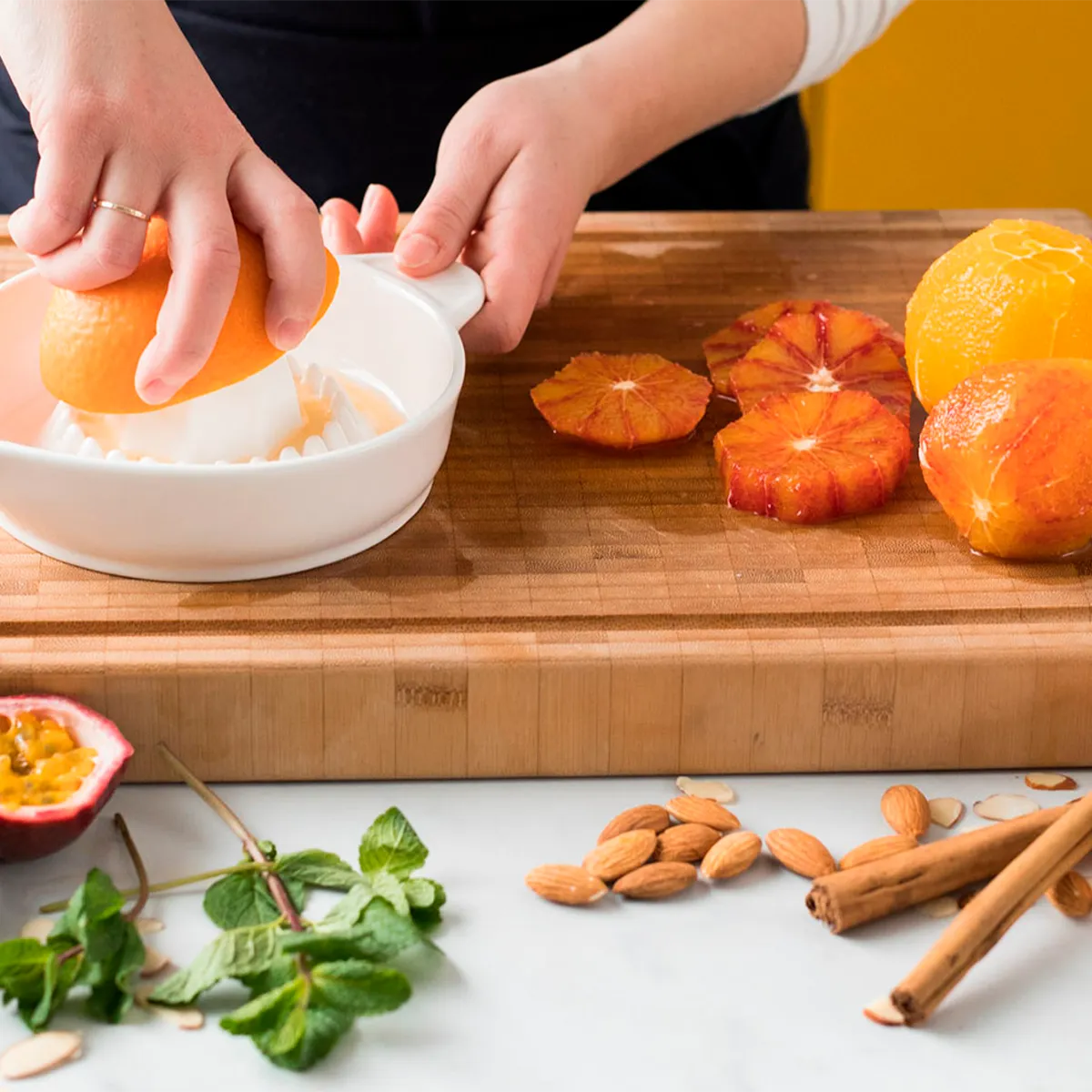 Person using a white citrus juicer with oranges, on a wooden cutting board with almonds and mint.