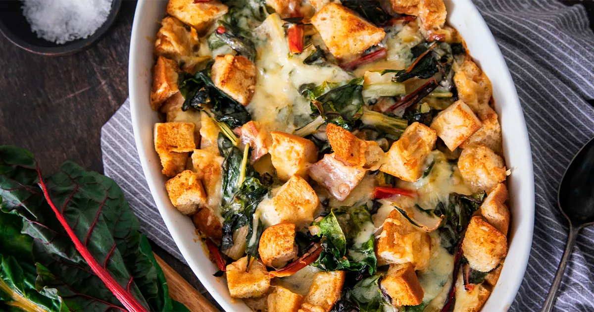 Baked casserole with bread cubes, cheese, and greens in an oval dish on a striped cloth.