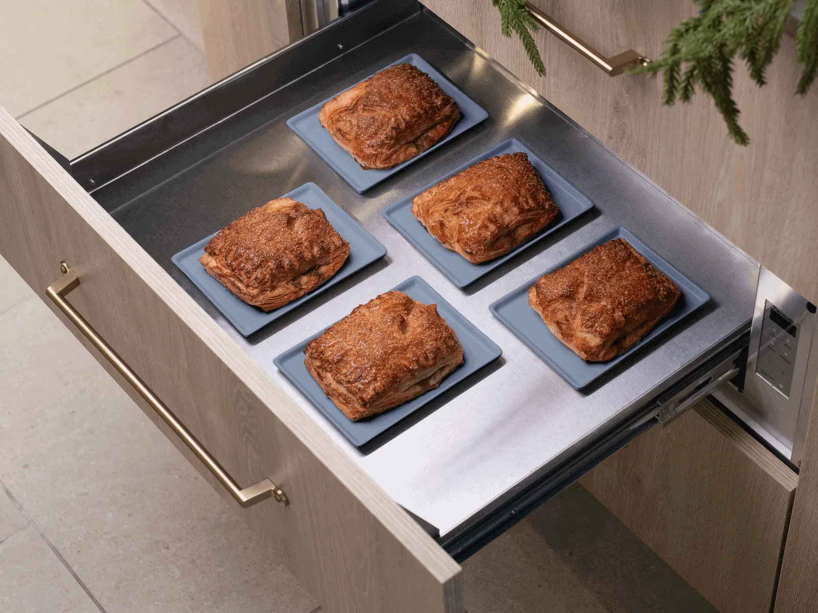An open Thermador warming drawer with a custom wood panel, showing five golden-brown pastries on individual plates inside.