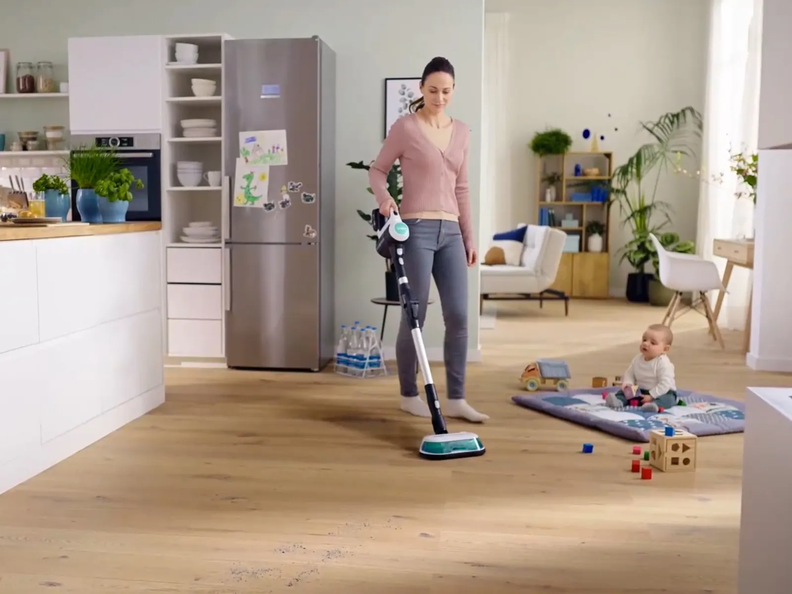 Woman vacuuming a mess on the floor in a bright kitchen while a baby sits on a play mat surrounded by toys.