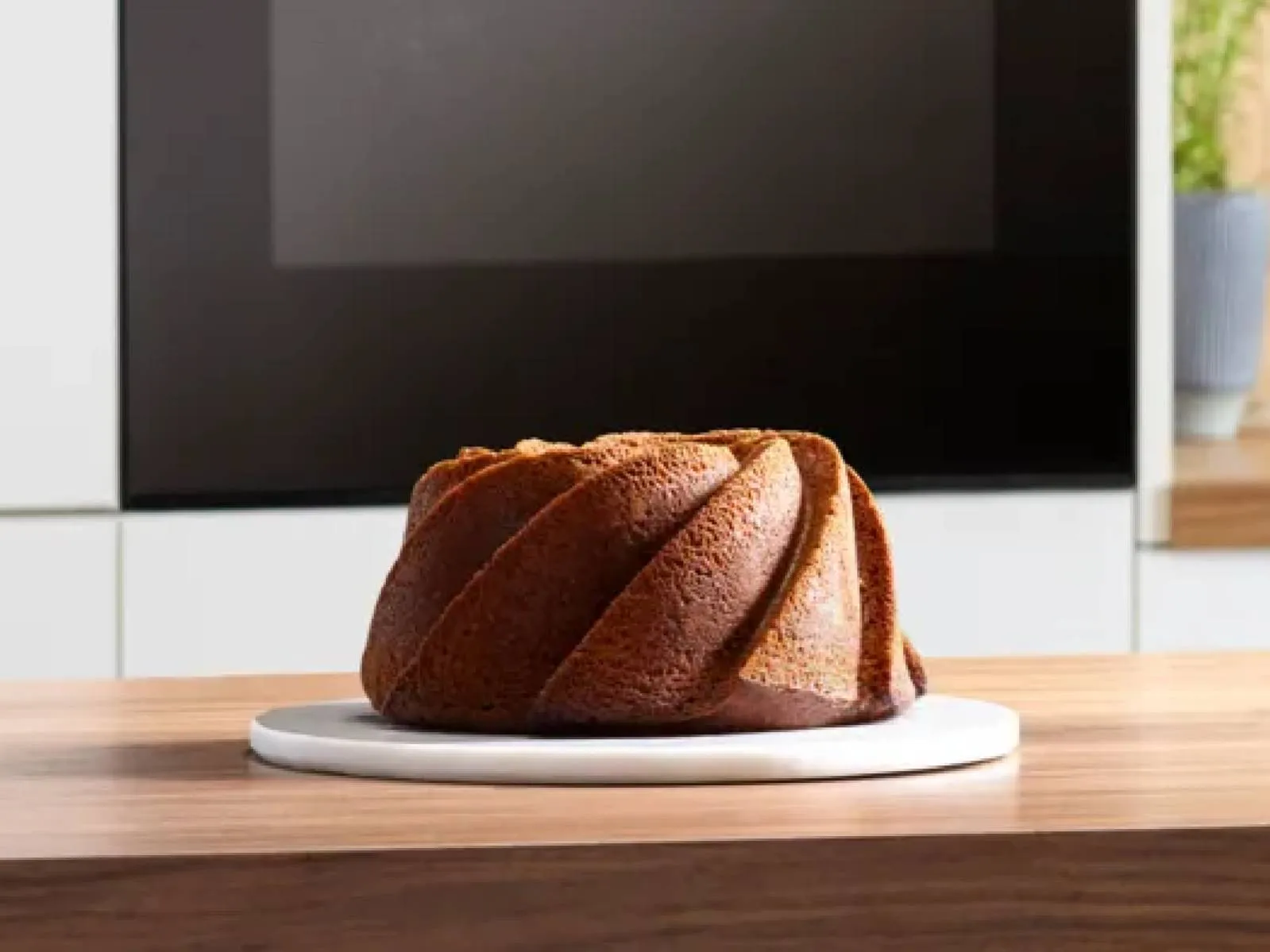A golden-brown bundt cake on a white plate graces a wooden table, set against a sleek modern kitchen backdrop.