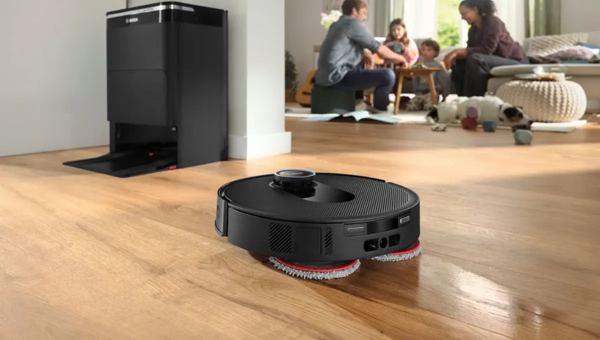 Robot usisavač i brisač čiste drveni pod, u blizini se nalazi kućna stanica za punjenje, a u pozadini se igra obitelj.