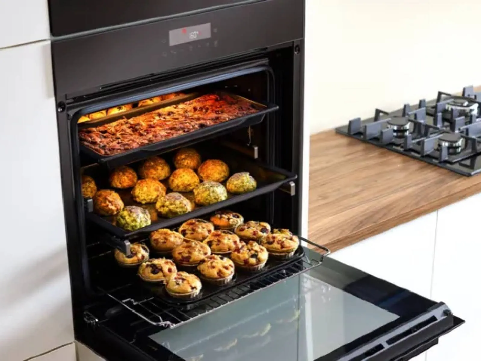 A modern black Bosch oven holds three trays of baked goods, including muffins and savory bites, against a warm wooden countertop.