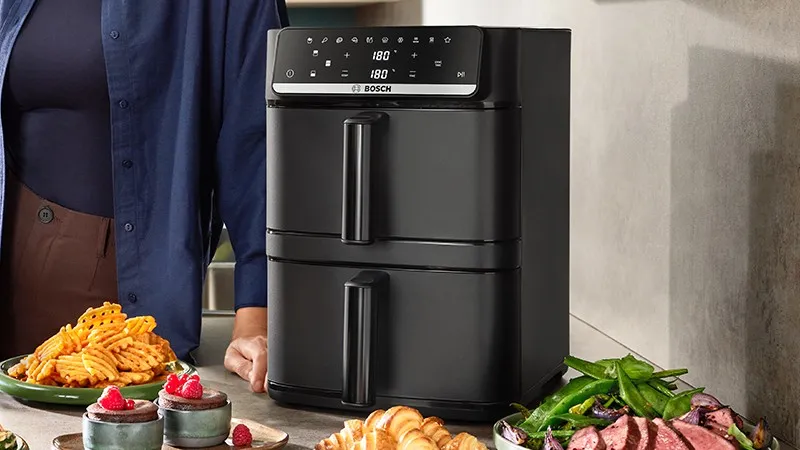 Bosch double drawer air fryer on a kitchen counter with waffle fries, desserts, roasted vegetables, and sliced meat nearby. 