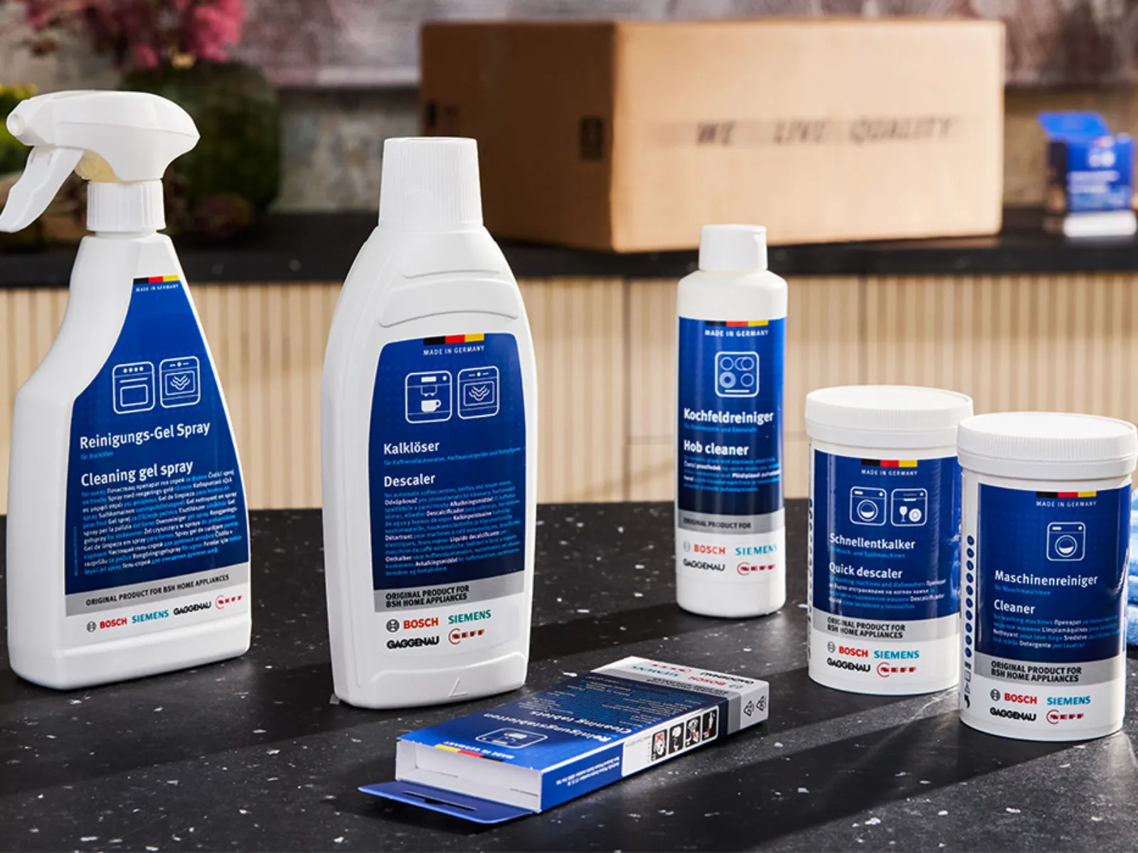 Several white bottles and tubs with blue labels — a cleaning gel spray, descaler, hob cleaner, quick descaler and machine cleaner — arranged on a dark kitchen countertop with a folded blue cloth and a cardboard box in the blurred background