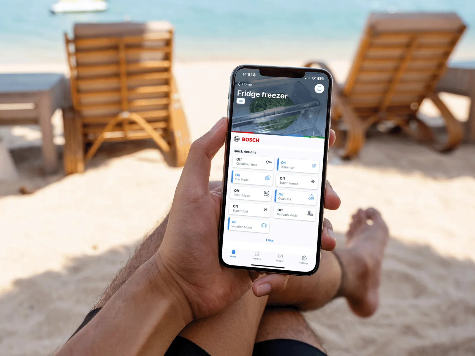 A person relaxes on a chair, using a smartphone to manage their Bosch fridge settings by the ocean.