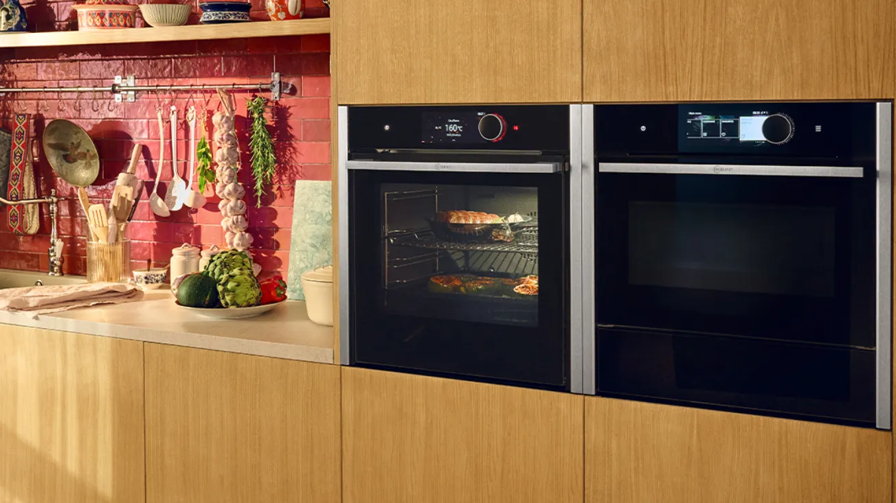 Kitchen counter with red tile backsplash, hanging utensils, garlic and herbs, and a plate of artichokes and vegetables; two black built‑in ovens with stainless trim — left oven illuminated showing food inside.