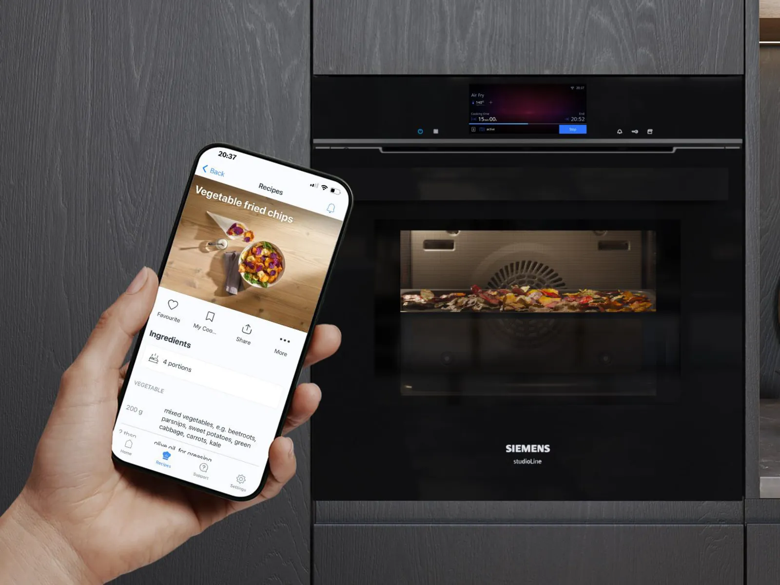 Une main tenant un téléphone qui affiche une recette de chips de légumes, à côté d'un four encastrable noir Siemens dans lequel cuisent différents légumes.