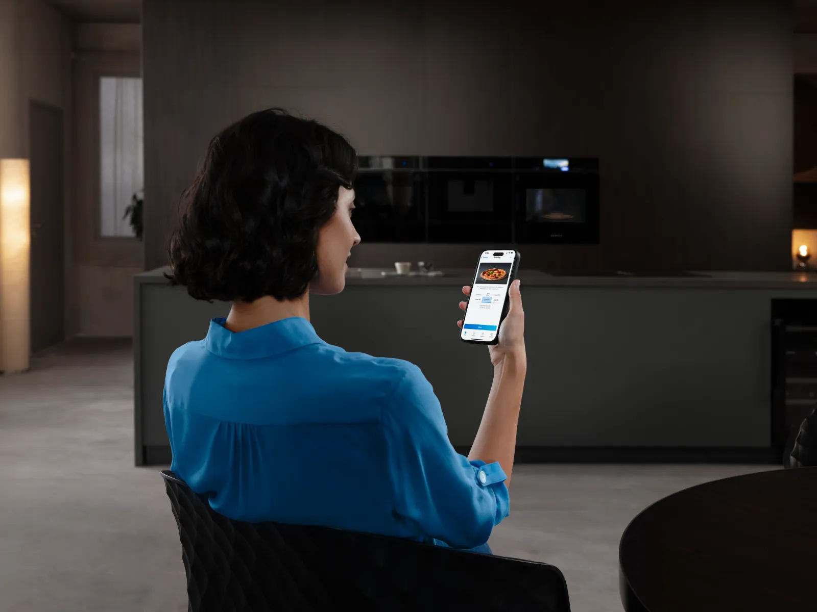 Kvinna i blå skjorta sitter i ett modernt kök och tittar på en smartphone som visar en matleveransapp med en pizza på skärmen.