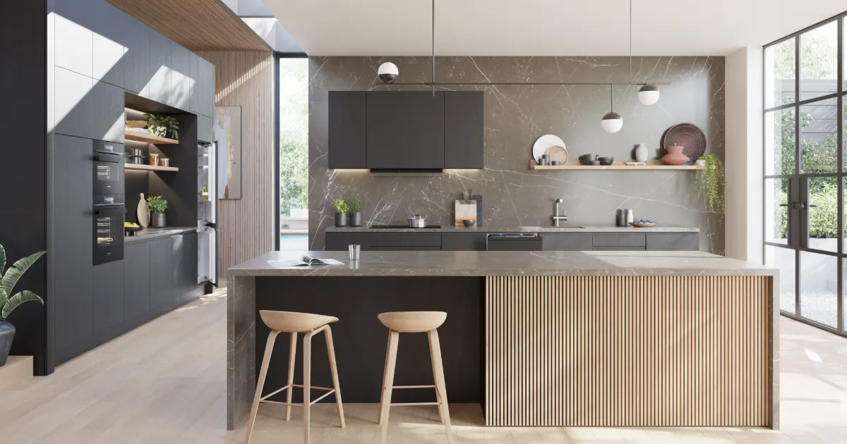 A wide view of a modern, high-end kitchen featuring dark grey cabinets, a grey marble backsplash, and a large island with a fluted wood base. The kitchen is equipped with built-in black ovens and an integrated refrigerator. Two wooden stools are tucked under the island, and the space is brightly lit by large windows and pendant lighting.