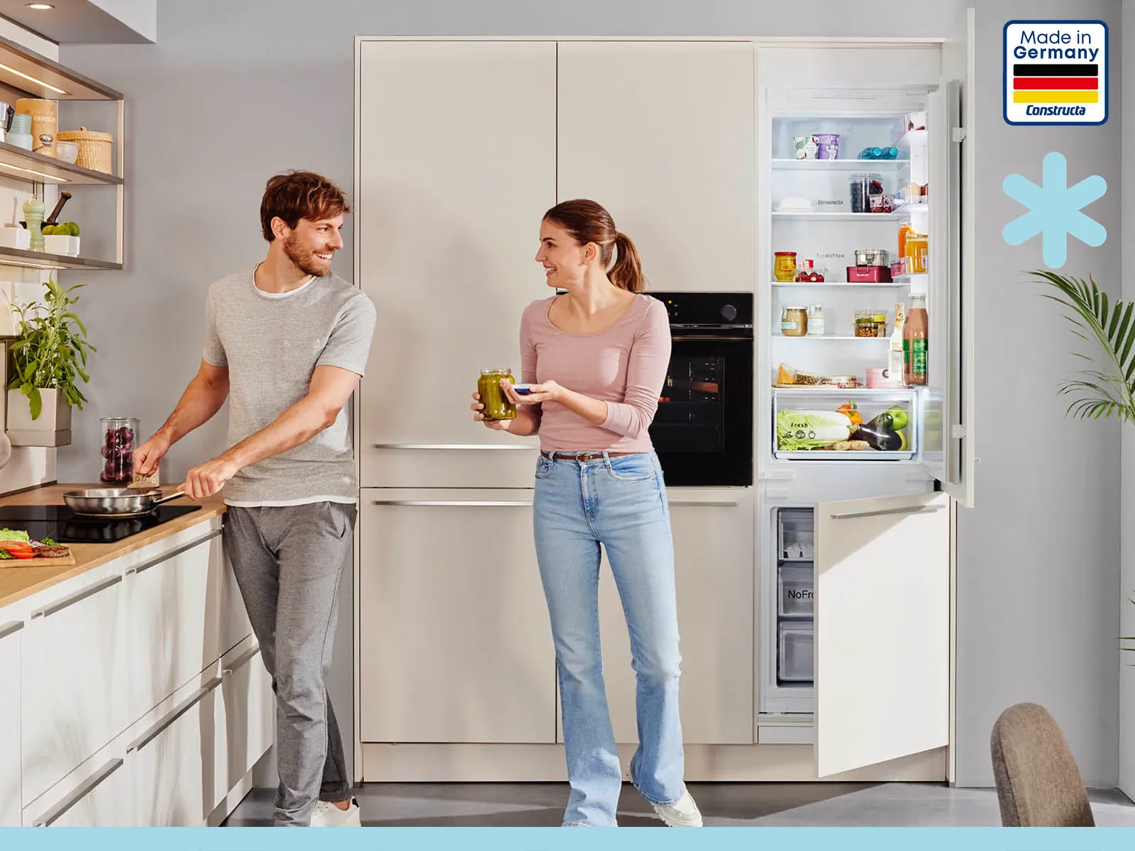 Ein junges Paar in einer modernen, hellen Küche. Der Mann kocht am Herd, während die Frau etwas aus dem geöffneten Constructa Einbau-Kühlschrank holt.