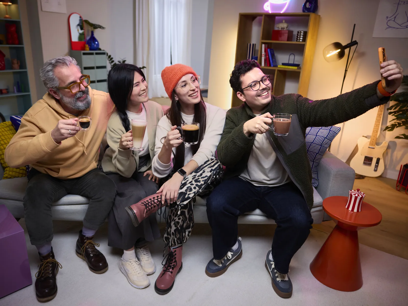 A group of four diverse friends enjoys coffee at home. They are sitting on a sofa, smiling and taking a selfie.