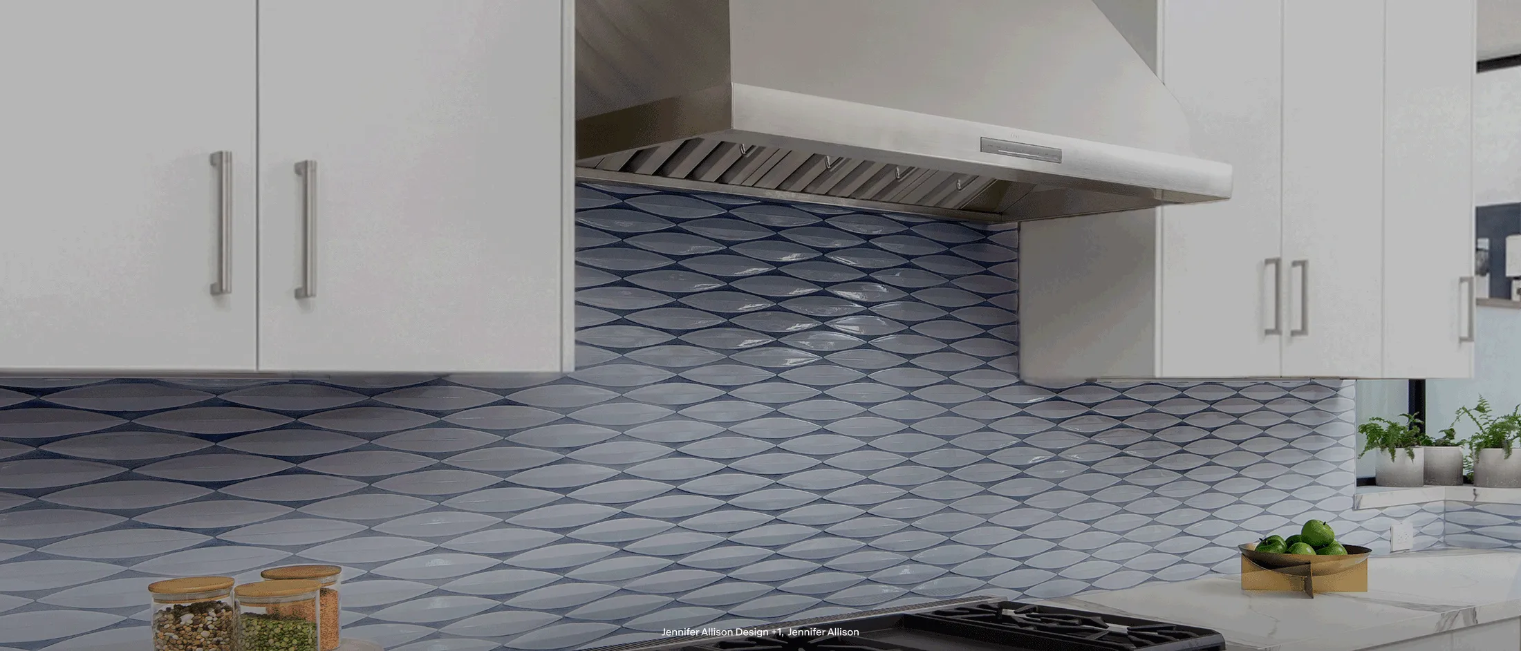 Modern kitchen with patterned blue and white tile backsplash, gas stove, wooden utensils, glass jars, and stainless steel fridge.