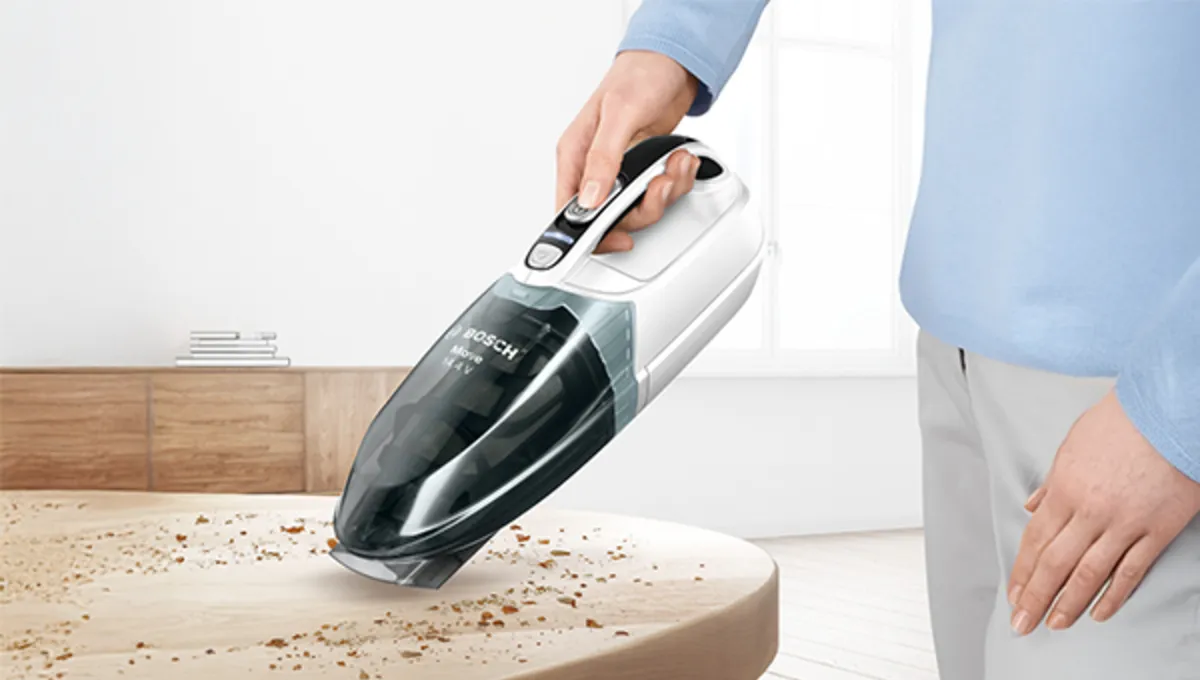A person uses a handheld vacuum cleaner to clean up crumbs from a wooden table in a bright, minimalist room.