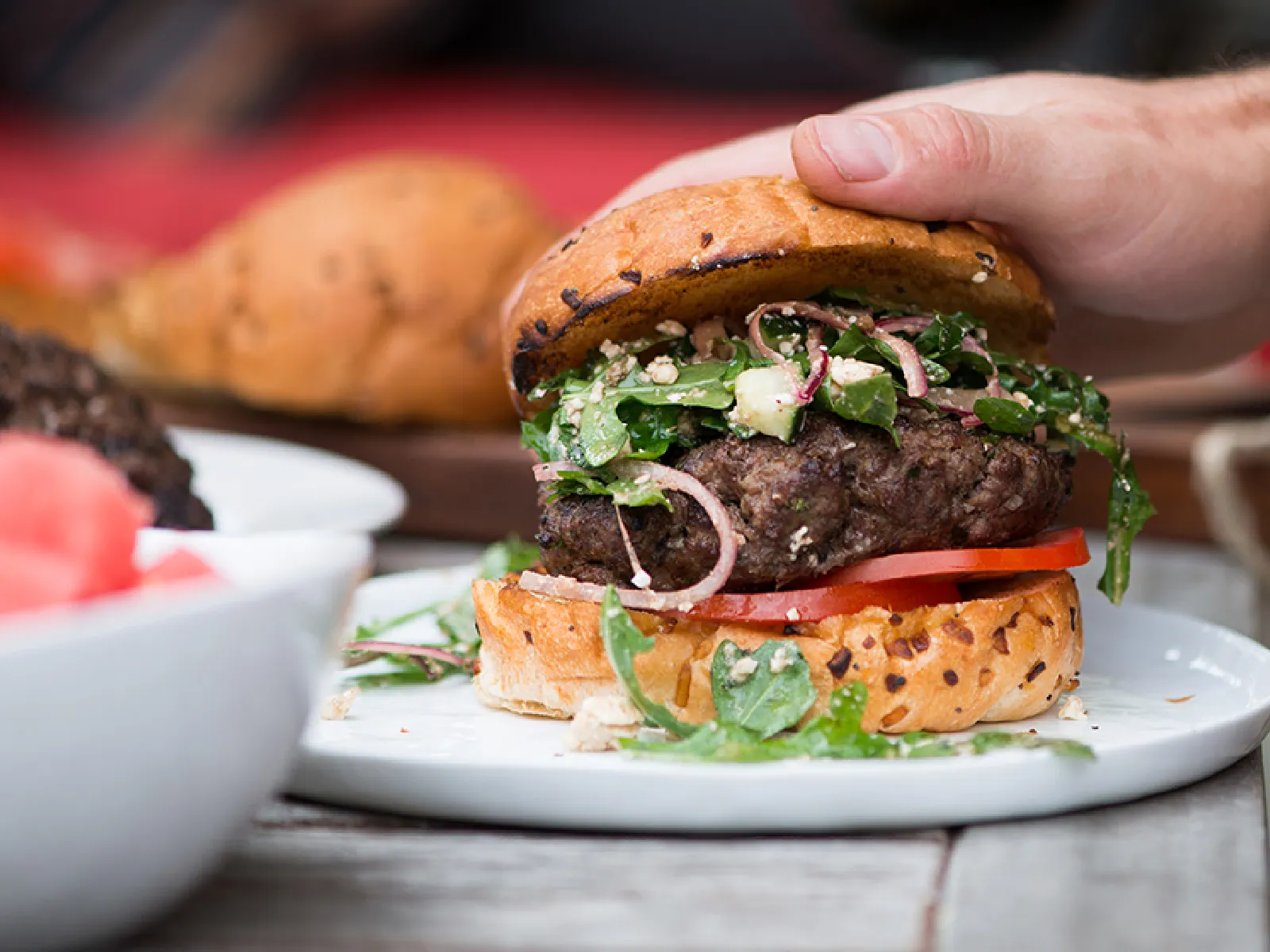 Hand holding a juicy burger with lettuce, tomato, onions, and sauce on a poppy seed bun served on a white plate.