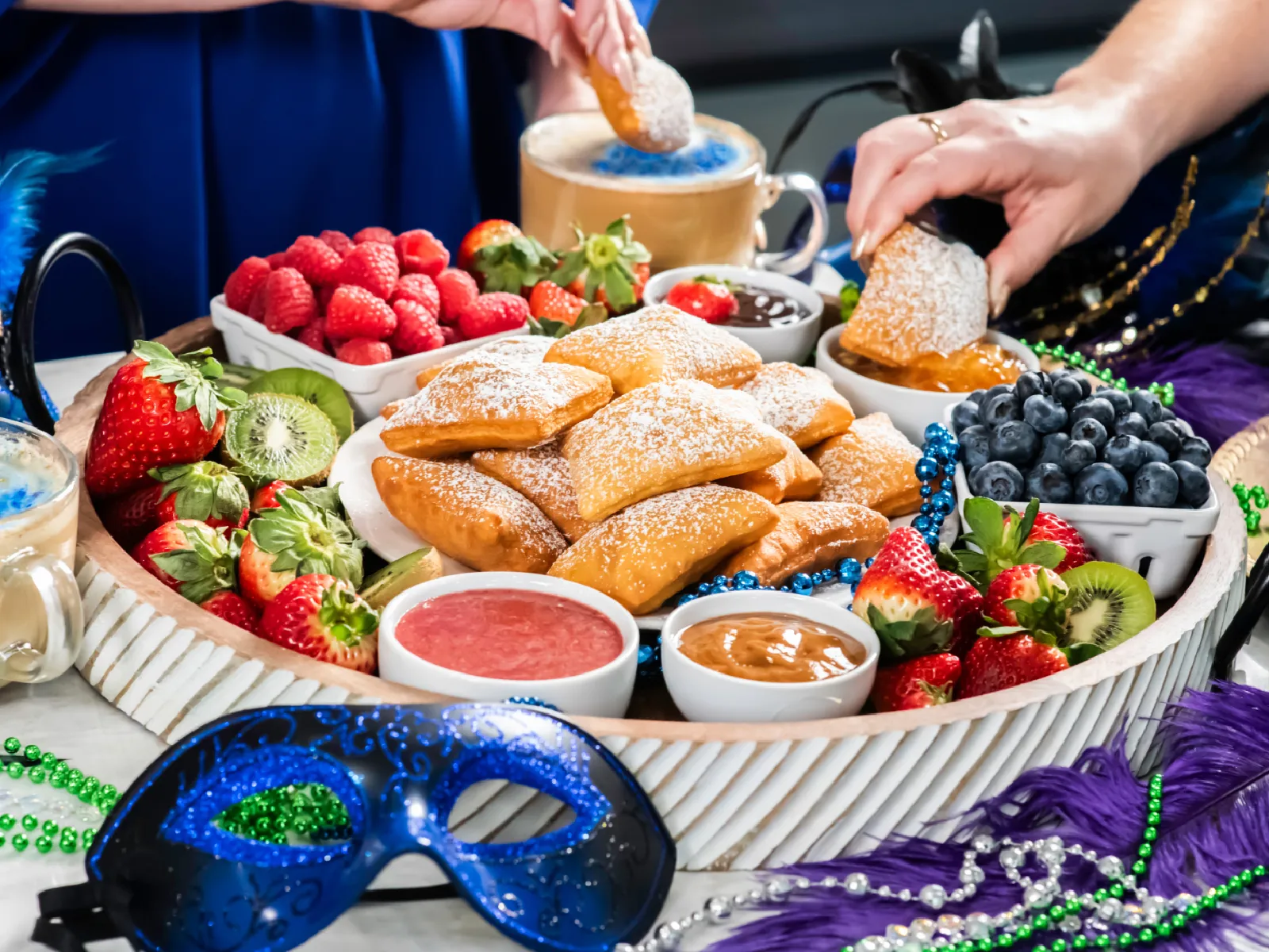Tray of beignets, fresh berries, and dipping sauces with coffee, Mardi Gras masks, and beads on the table during a festive gathering.
