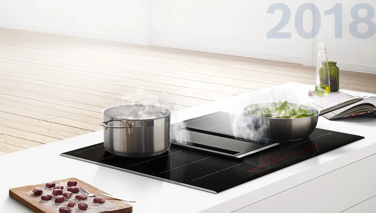 A Bosch induction hob with integrated downdraft ventilation on a white kitchen island, with two steaming stainless steel pots and fresh ingredients on a nearby chopping board, 2018.